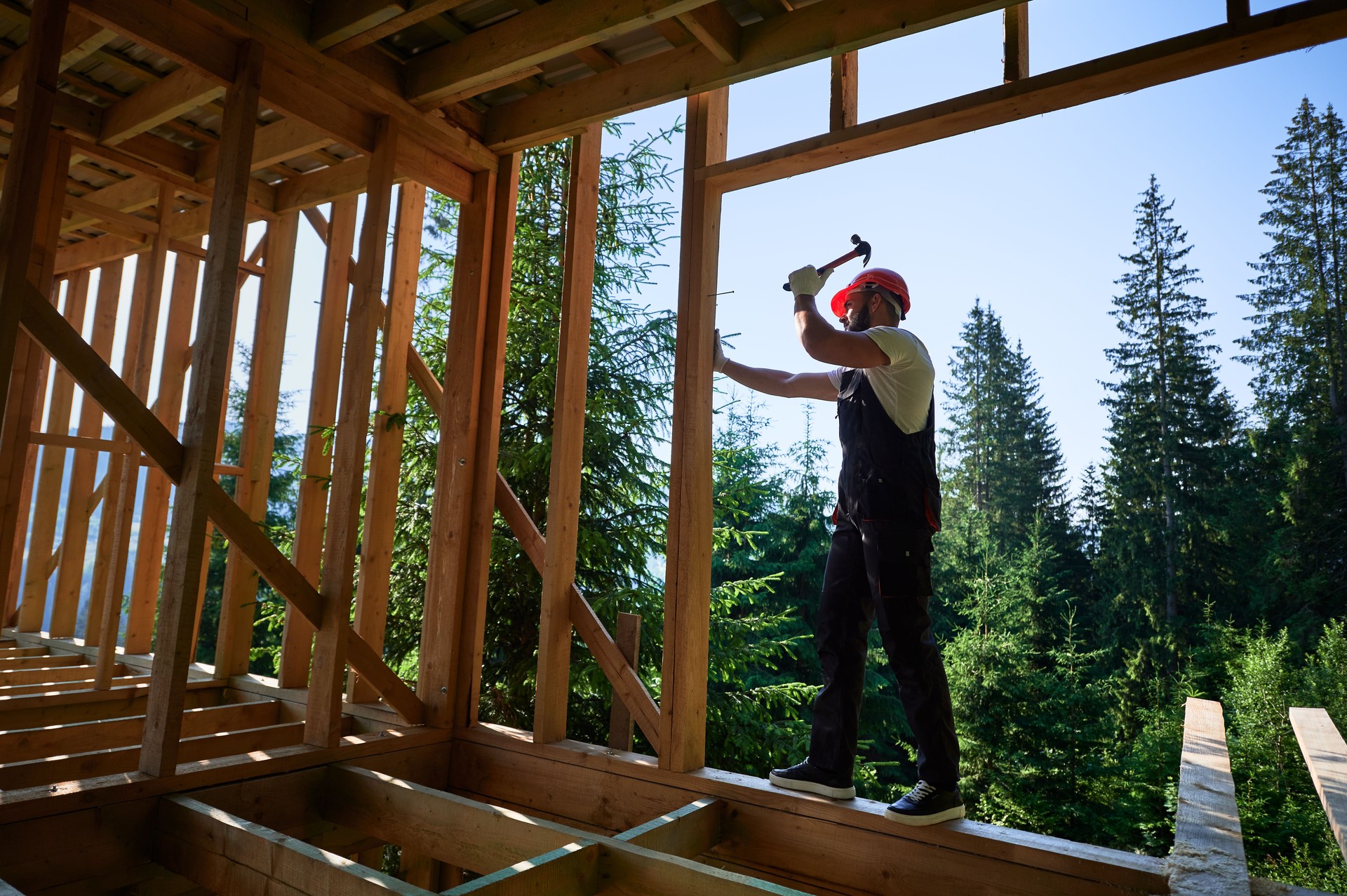 Carpenter hammering nail while constructing wooden frame two-story house near the forest.